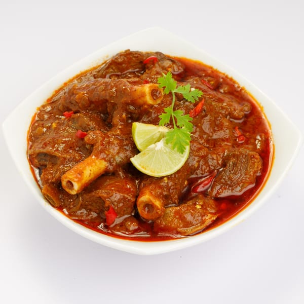 Indian lamb curry with bone-in meat, garnished with lime wedges and cilantro in a white bowl.