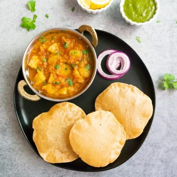 Aloo curry with puri bread, red onion, lemon, and green chutney on a black plate.