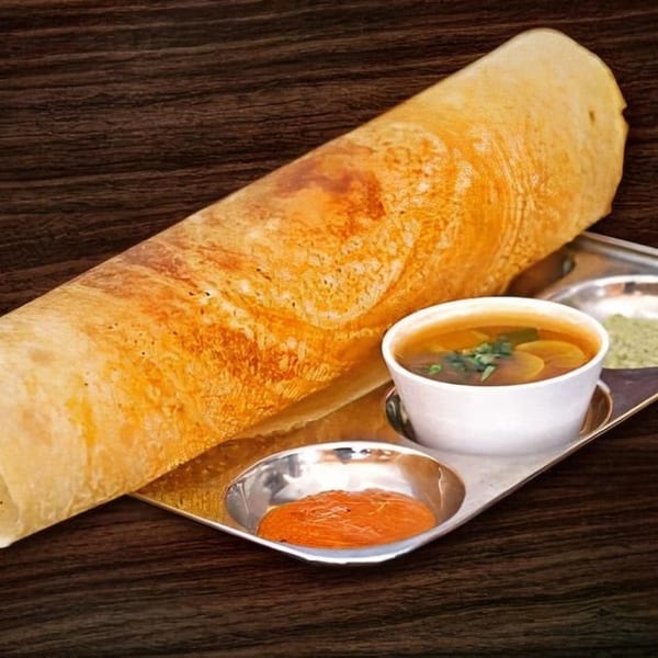 A golden-brown dosa, rolled and served on a silver tray with two chutneys and a bowl of sambar.