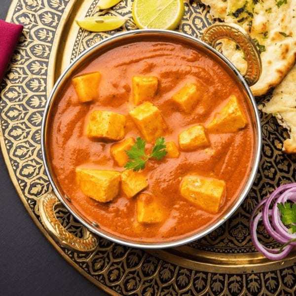 Paneer butter masala in a brass bowl with naan, lemon, and red onion on a decorative tray.