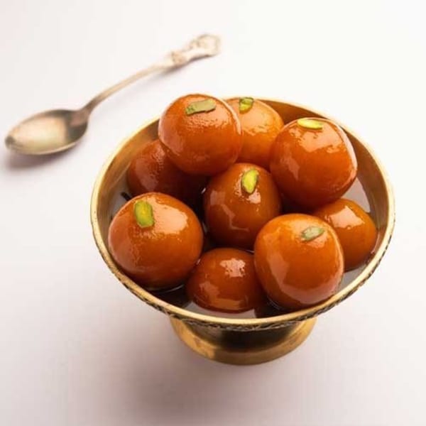 A brass bowl filled with glistening Gulab Jamun, garnished with pistachios, and a spoon nearby.